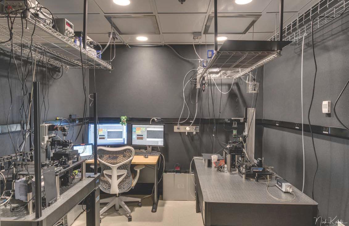 view of room from back with 3 computer screens on a desk, with a chair facing forward. Machines lines walls on left and right and wires are running from ceiling to electrical outlets beside the computer screens.