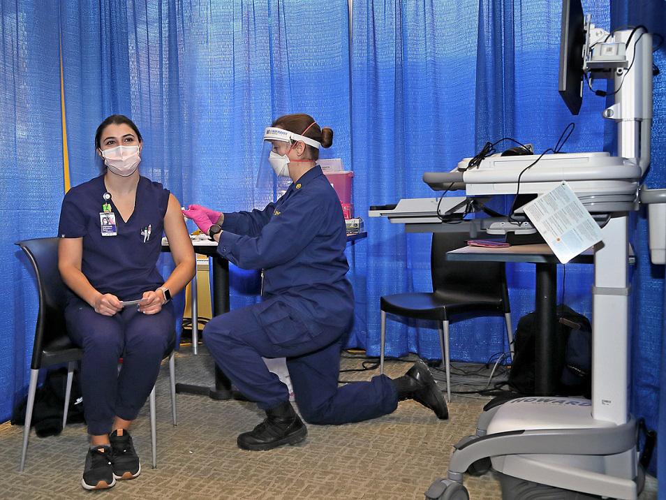 A masked who is also wearing a face shield gives another masked nurse a vaccination.