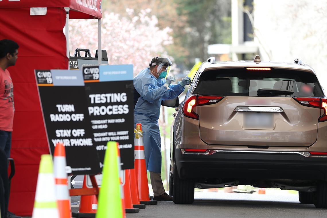 A health professional wearing full PPE speaks to someone sitting in a midsize SUV.