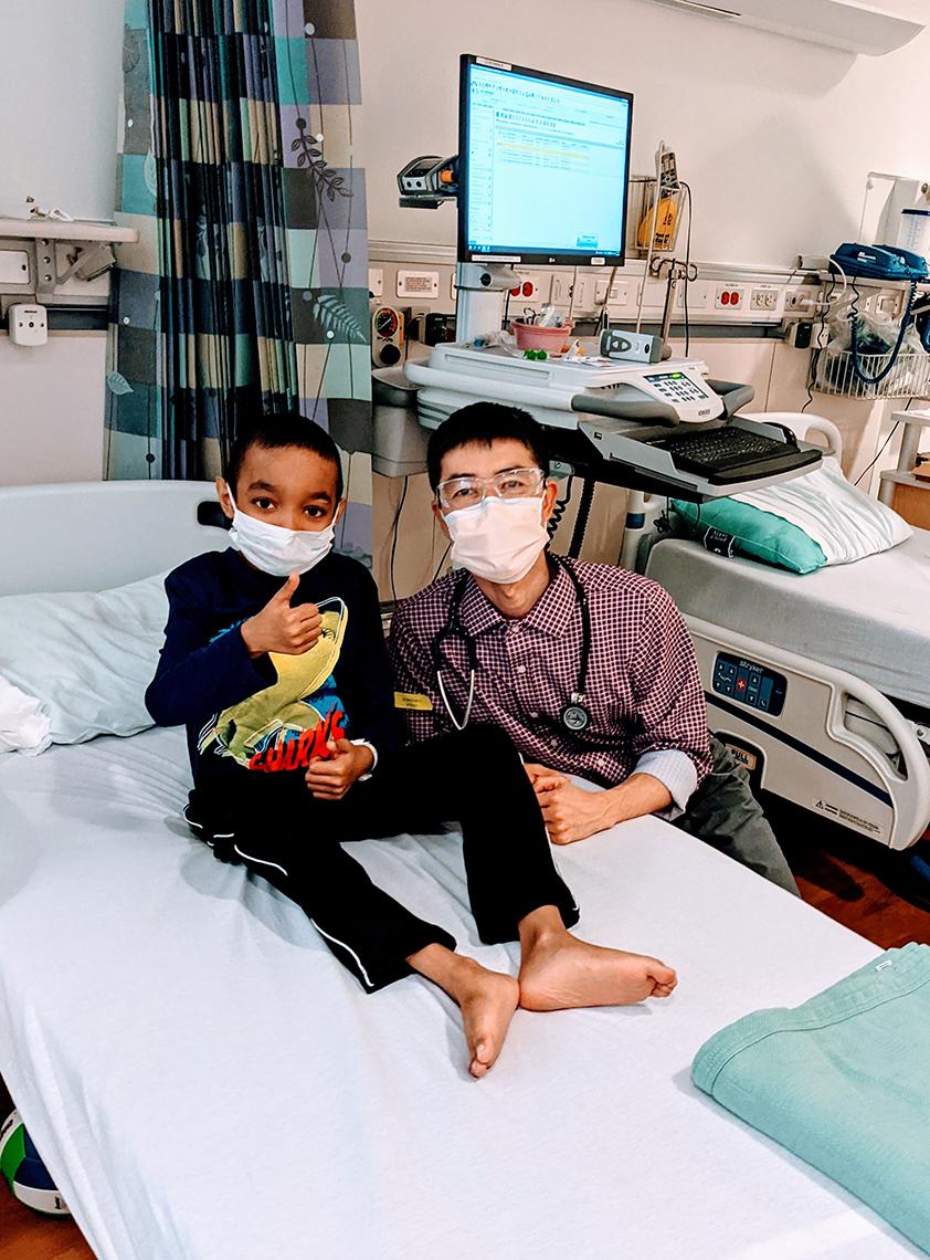 Sant sits up in his hospital bed giving a thumbs up with Dr. Hsieh (pronounced "Shay") kneeling by the bed.