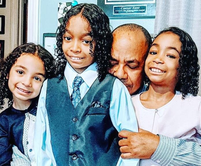Father Lucas Sant, solemn with eyes closed, hugs his 3 smiling children