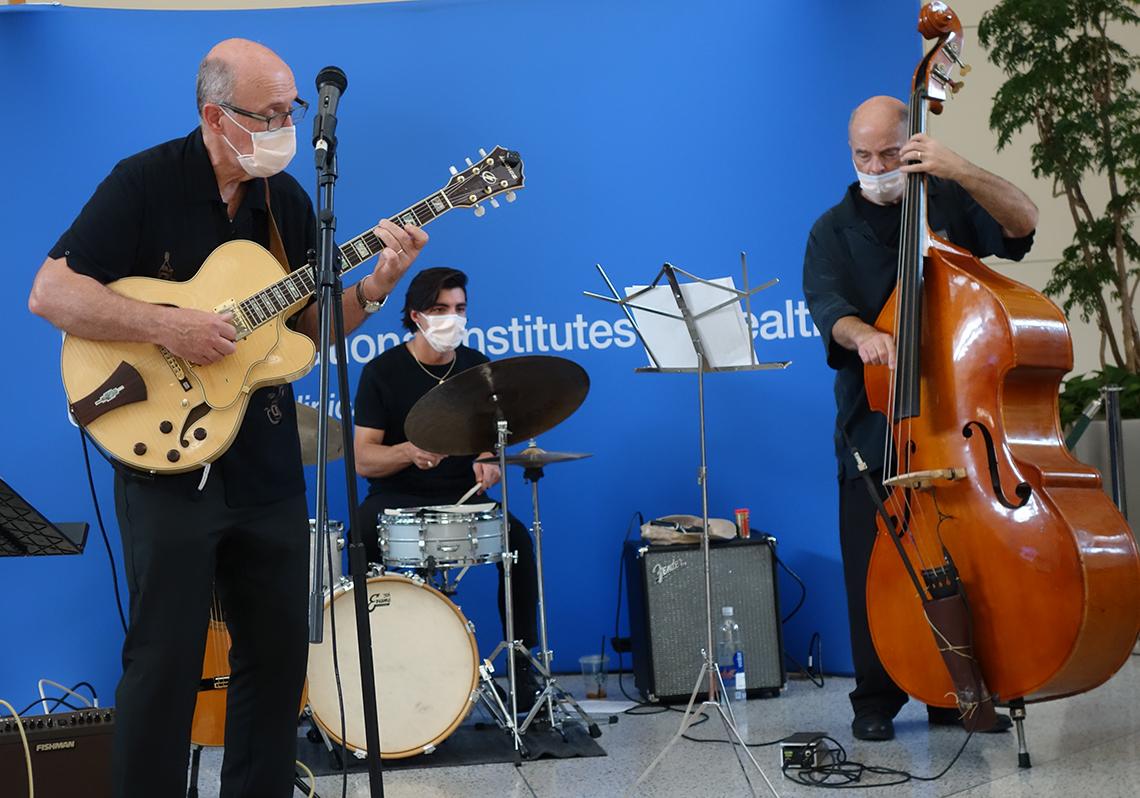 Three men in the CC atrium, playing guitar, drums and bass