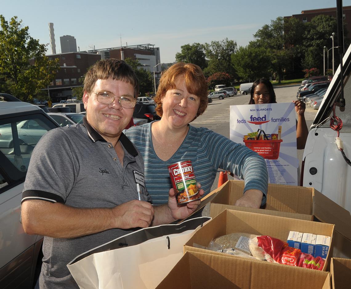 A man holds a can of Chunky Beef soup while a woman puts an object in a cardboard box full of nonperishable food.