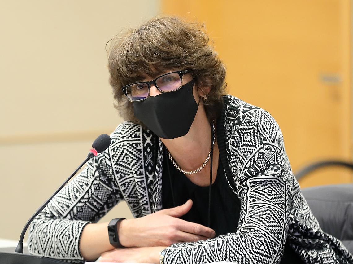 Erbelding, in black mask with arms folded, sitting in front of yellow wall, speaks.