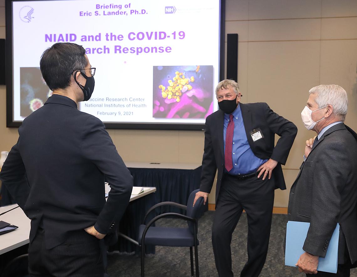 Koizumi, Lander and Fauci stand talking in front of video screen with purple slide, "NIAID and the COVID -19 Research Response"