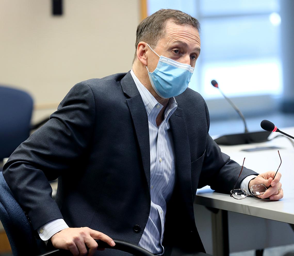 Mascola, in blue mask, sits holding eyeglasses.