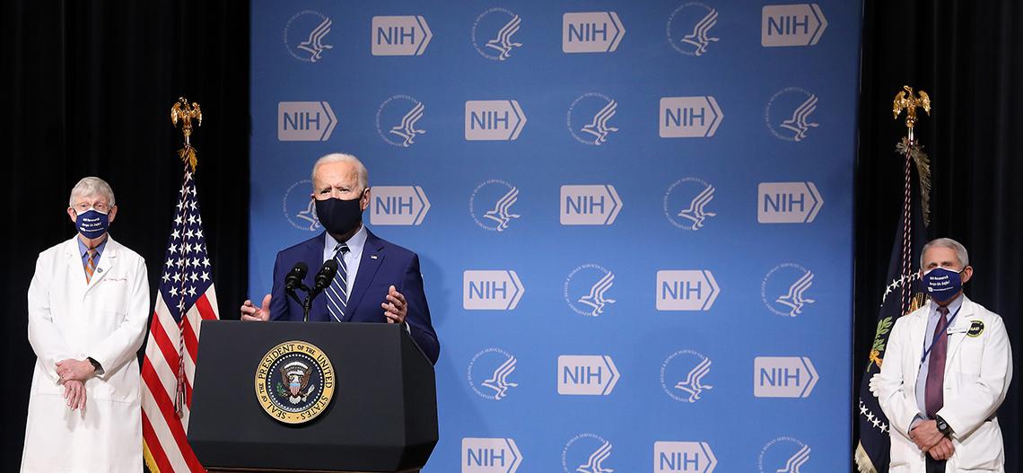 Flanked by Collins (l) and Fauci (r) on stage, Biden speaks from podium.