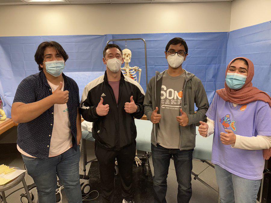 Four masked students gives a thumb up hand gesture. Behind them is a model skeleton.