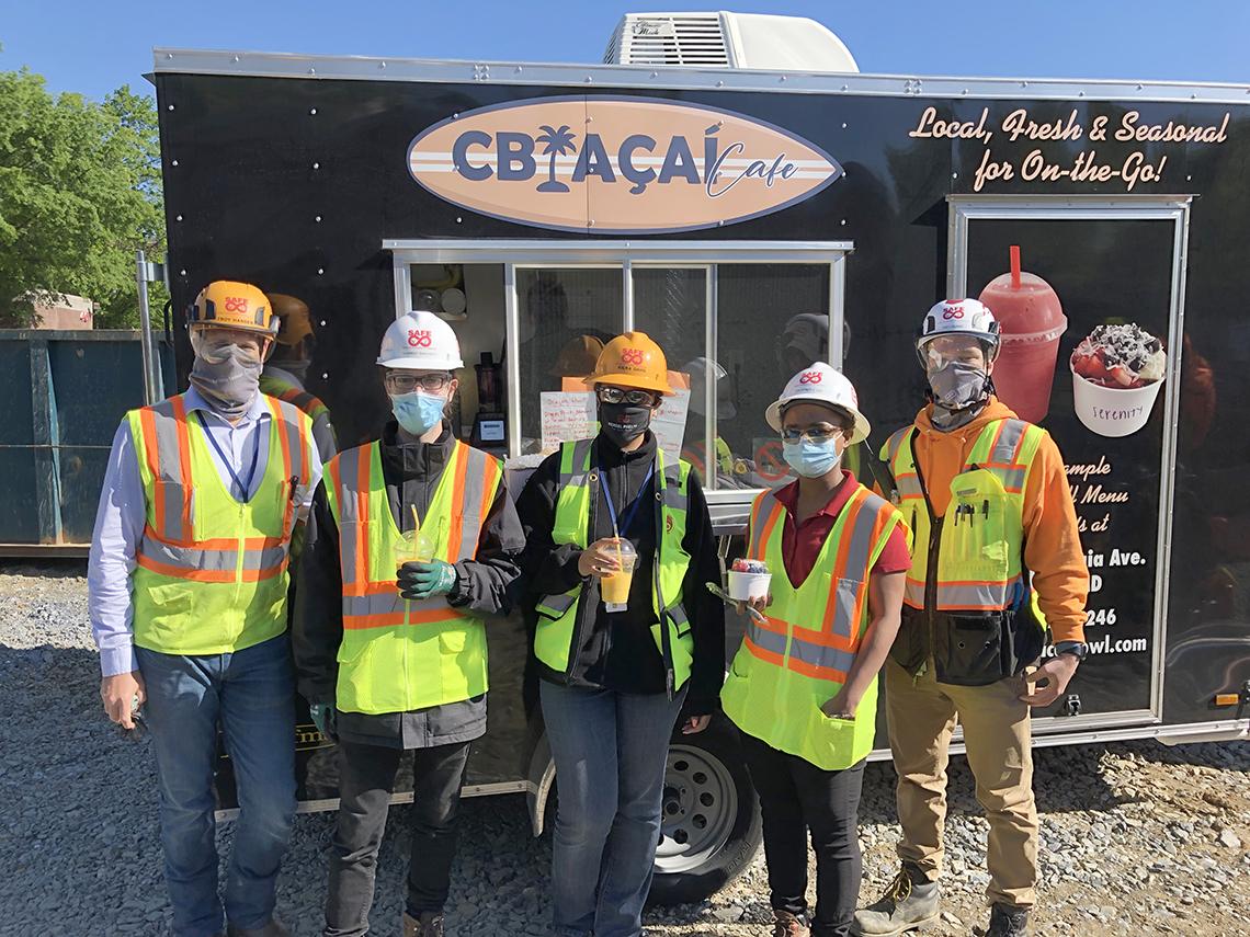 Construction workers in safety helmets and vests smile in front a food truck.