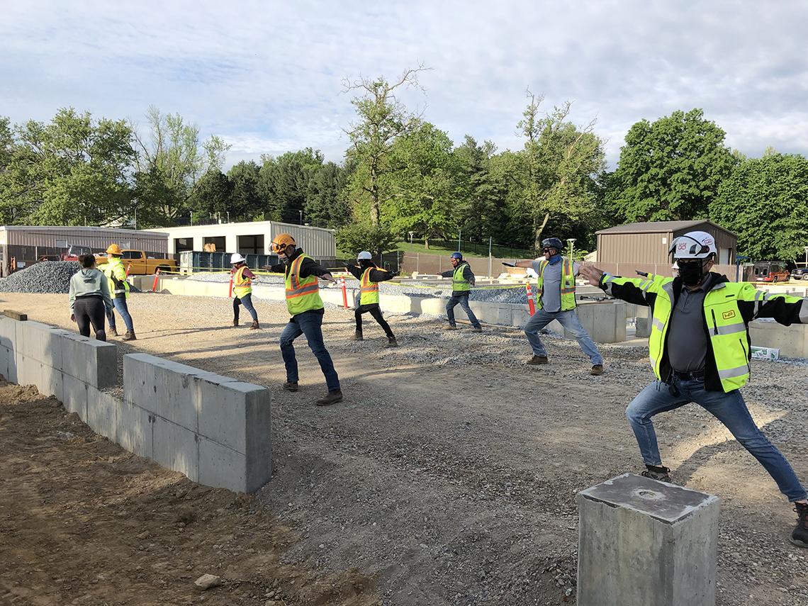 Instructor gesturing toward group of construction crew members who are posing with arms outstretched.