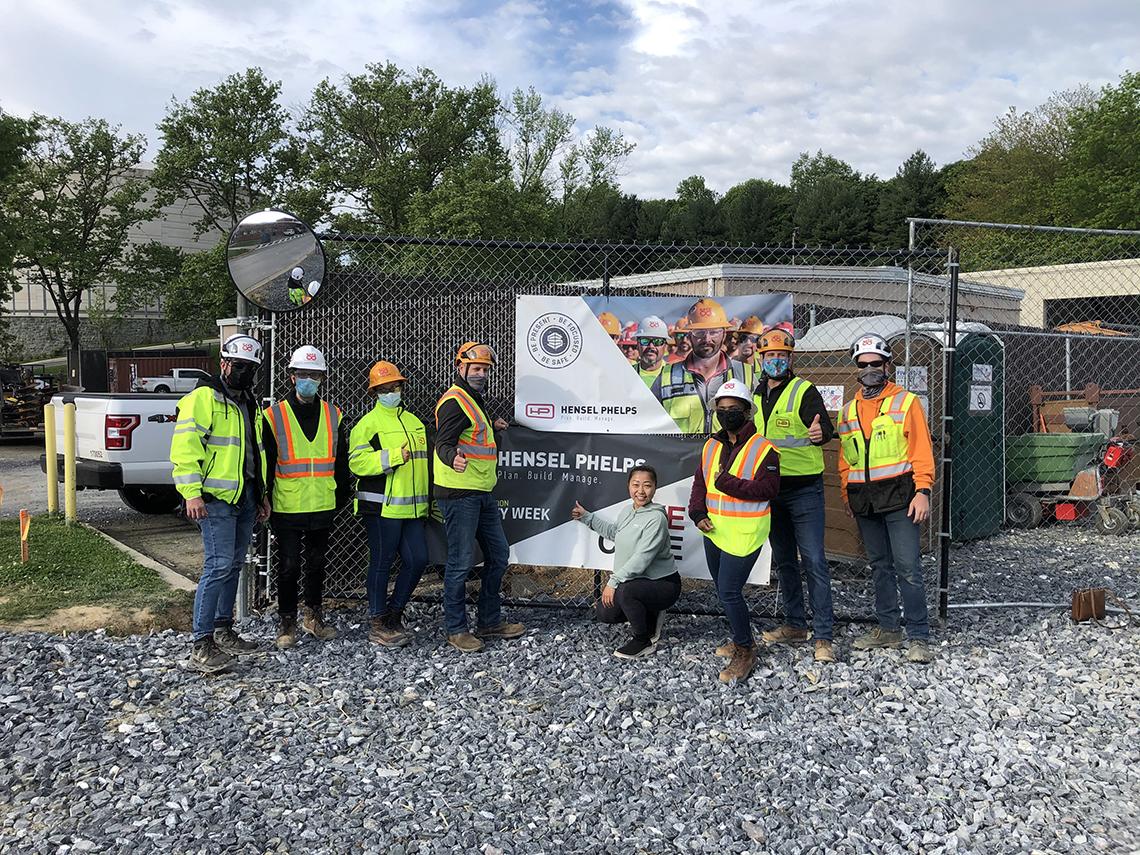 Several construction workers in safety helmets and vests pose at job site with a yoga instructor in front of company sign.