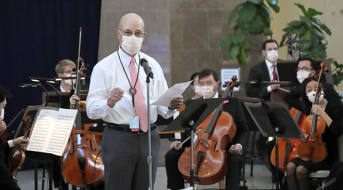 Gilman stands at microphone near musician holding his cello in the atrium.