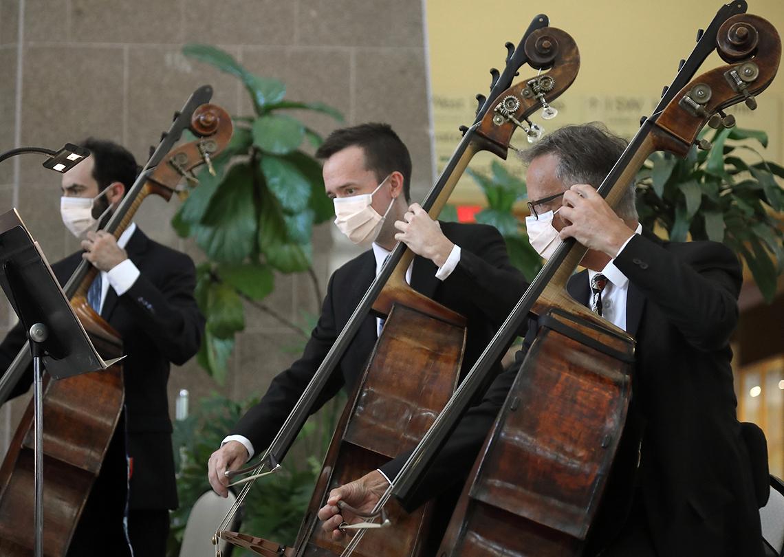 Three men wearing masks glide their bows across their cellos.