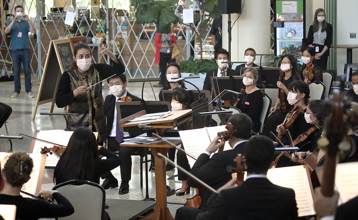 Members of the orchestra, all in masks with their string instruments and sheet music, follow the conductor as several staff members and a doctor in blue scrubs look on.