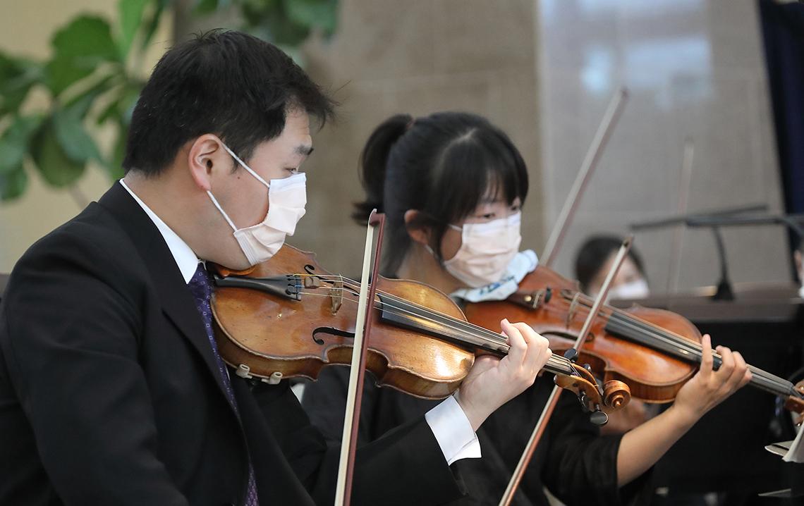 Two Asian musicians, wearing masks, hold their bows up to their violins in the atrium.