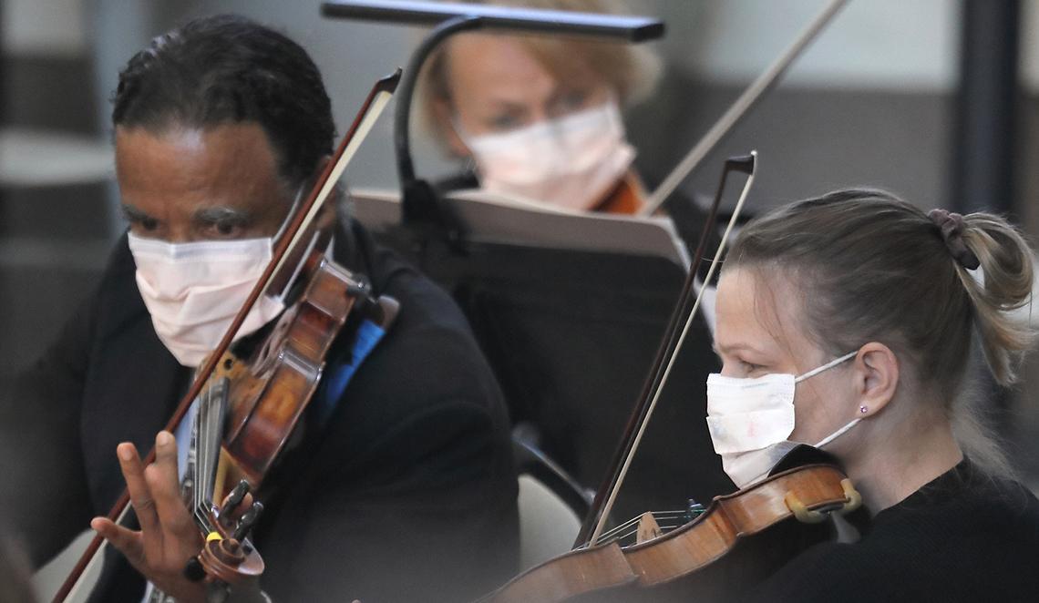Three masked musicians look toward music stands playing their violins.