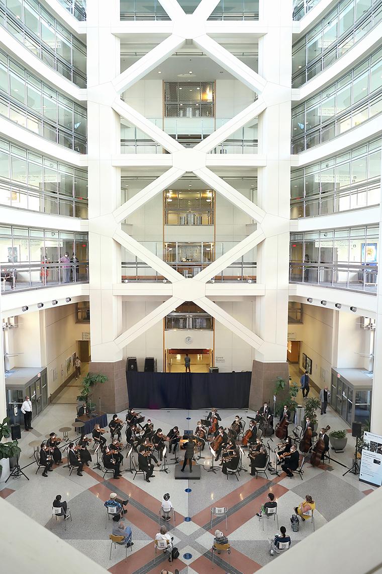 Aerial view of the atrium: orchestra performs with audience scattered, sitting 6 feet apart. Staff can be seen watching through windows in hallways above.
