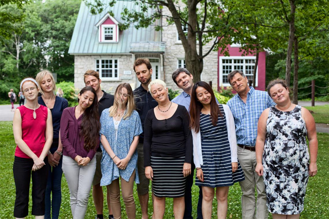 Charles Morin and his lab mates stand outside in front of colorful buildings, heads leaning to one side, all eyes closed, pretending to sleep.