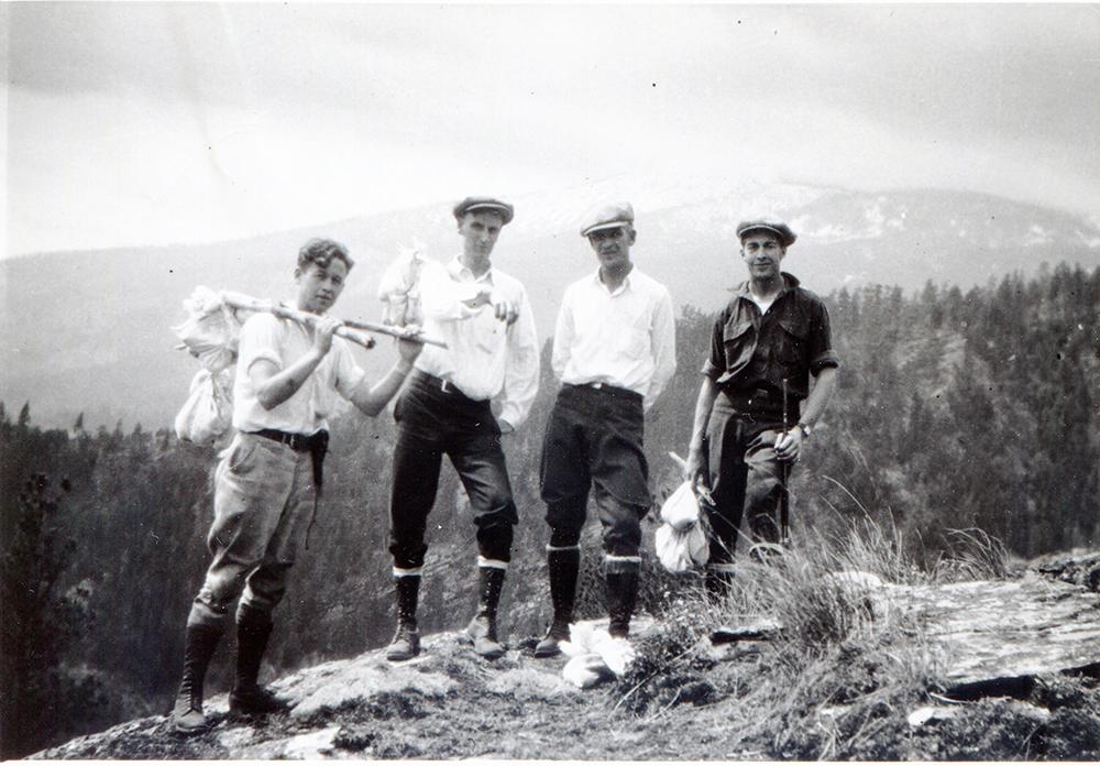 A black and white photo featuring four men holds several white cloth bundles tied to a stick for specimen collection. The background features a large snow covered mountain.