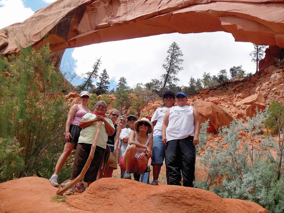 Research team members get a group photo taken below a red stone arch. Pine trees dot the area.