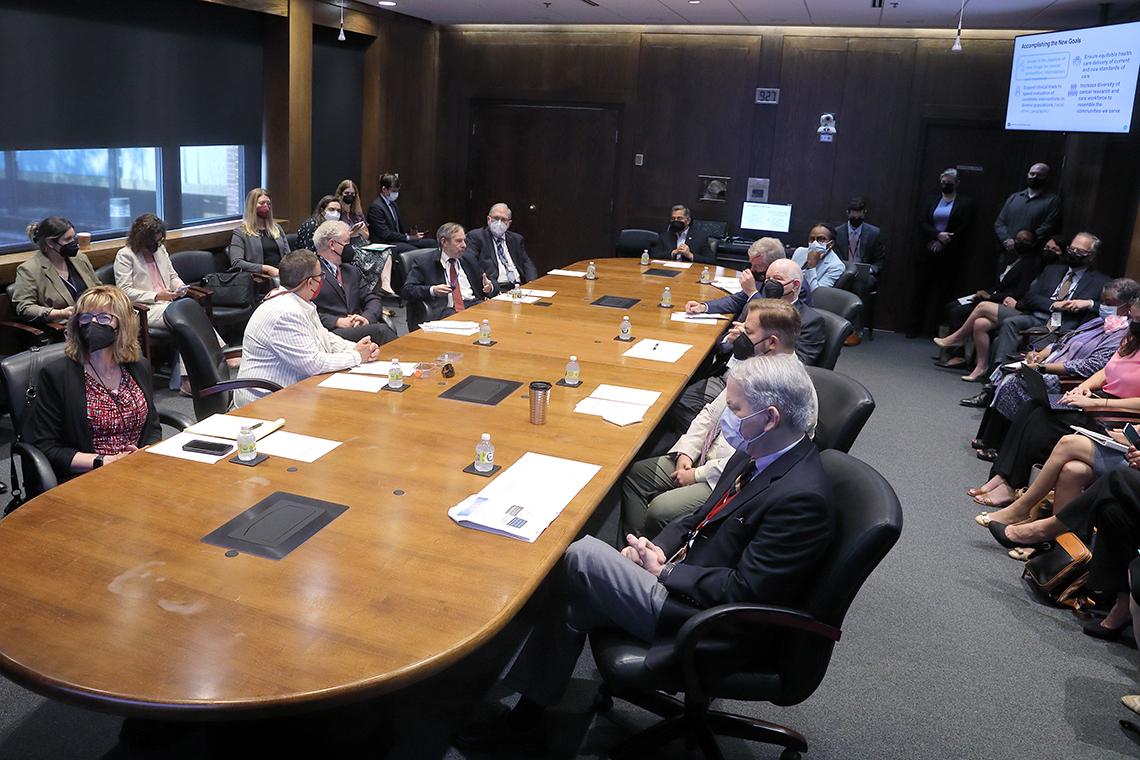 Conference room with people in masks seated around table and in chairs along the perimeter