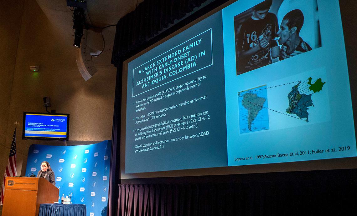 Quiroz stands at podium. Beside her, a slide of a boy feeding his father and a map of South America. The slide reads: A large extended family with early-onset Alzheimer's Disease in Antioquia, Colombia.