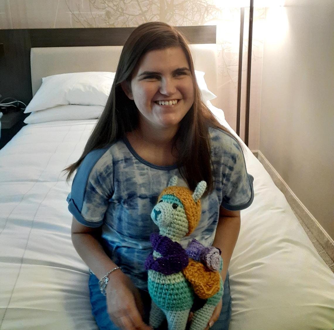 Sienna, with huge smile, sits on a bed holding her sheep plushie that's wearing a crocheted hat.