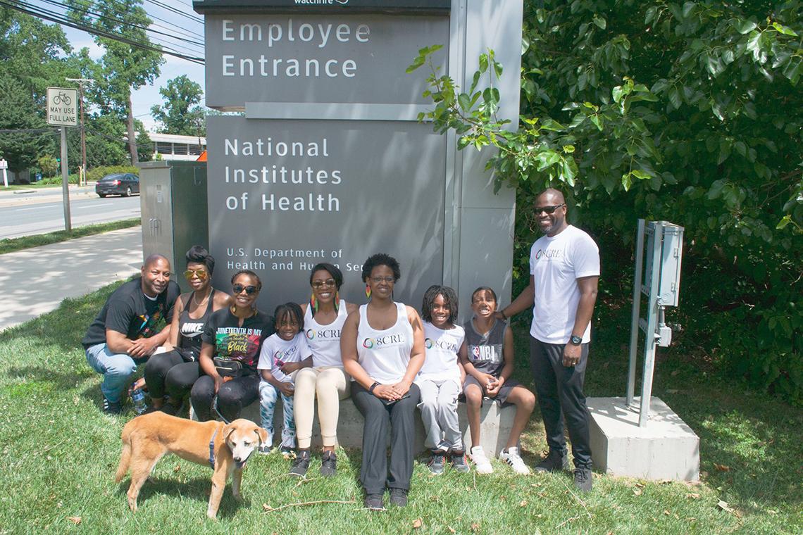 Group of adults and children in workout clothes smile in front of main NIH campus employee entrance sign