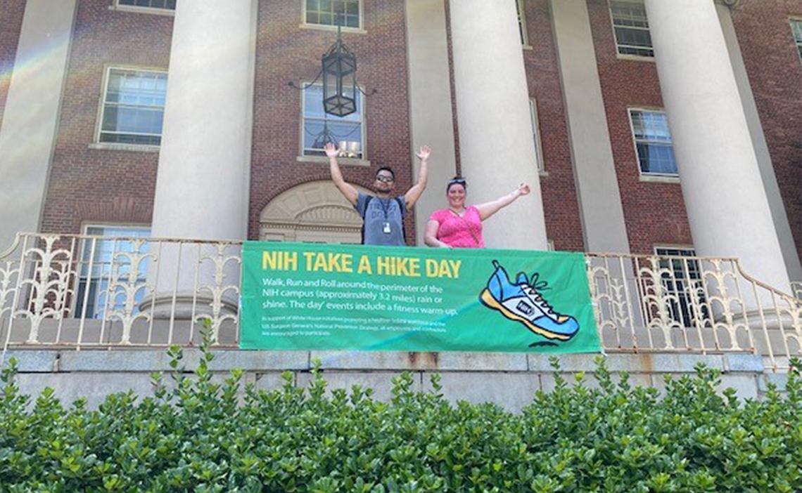 Castro and Younsi stand with arms high in the air in front of NIH Take a Hike Day banner outside Bldg. 1