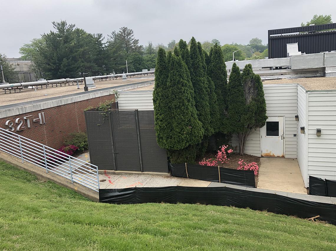 A view down the hill to a small brick building, 32T-II, flanked by tall trees, pink flowers and construction fencing