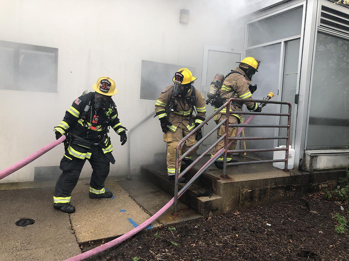 Three firefighters in full gear drag a hose up an outdoor staircase.
