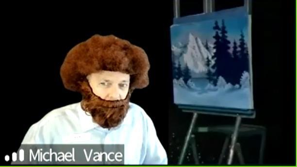 Head and shoulders of man wearing large brown curly wig, beard and mustache with landscape painting on easel behind him