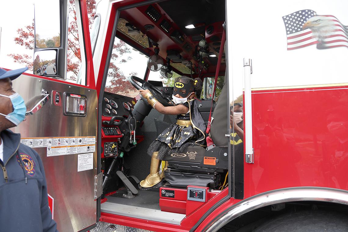 Youngster dressed as Batgirl sits behind wheel of fire engine.