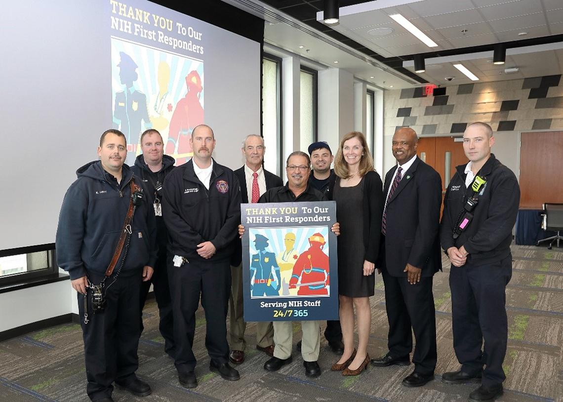Large group of people stand behind a poster with a graphic representing a firefighter, EMT, and police