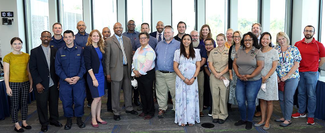 A large group of DOHS speakers and meeting attendees stand in conference room, posing together.