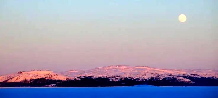Moon rises over blue water with snow-topped mountains in the distance