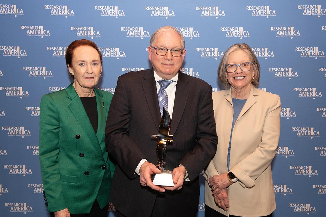 Mary Woolley and Ann Lurie stand with Dr. Tabak, who is holding award, against Research!America backdrop.