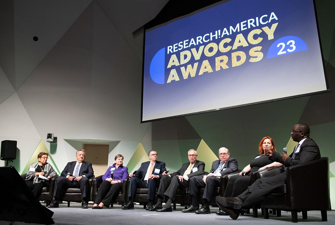 The 7 panelists sitting on stage listening to Dr. Olajide Williams, who is talking into microphone