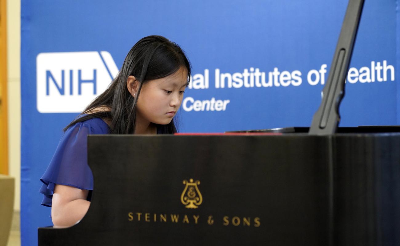 A young woman looks down at piano keys, with blue NIH backdrop.