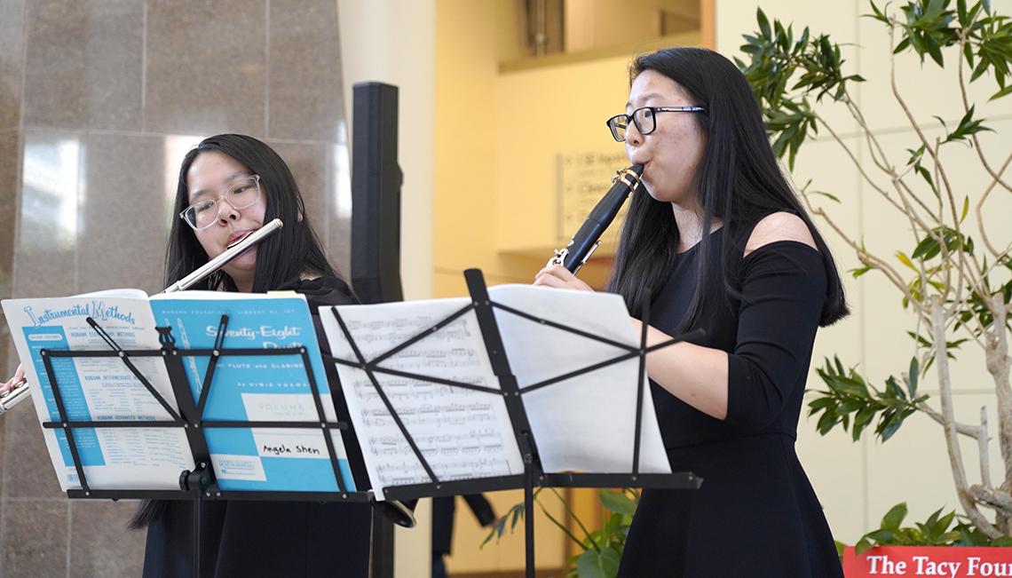 Two seated women, one playing flute and the other playing clarinet, looking at their music stands.