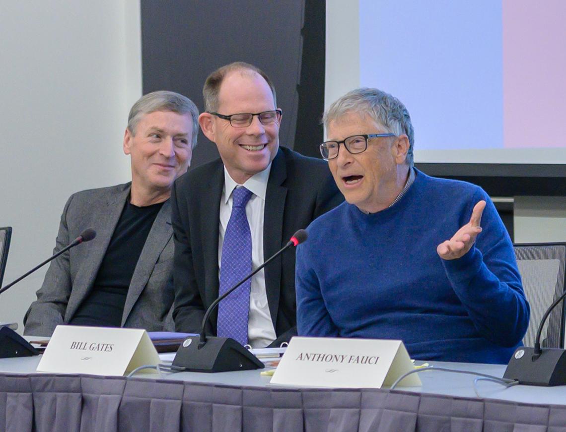 Gates speaks at a microphone while two men in suit jackets look on smiling