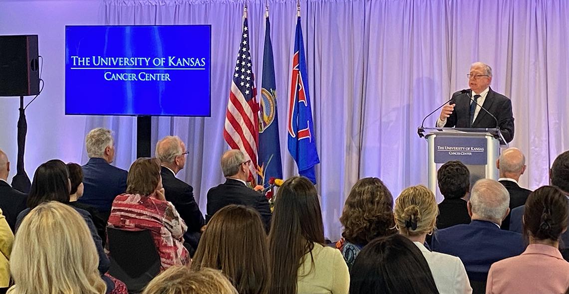A large assembled group sits in an auditorium listening to speaker at podium, with University of Kansas Cancer Center displayed on screen, flags on stage