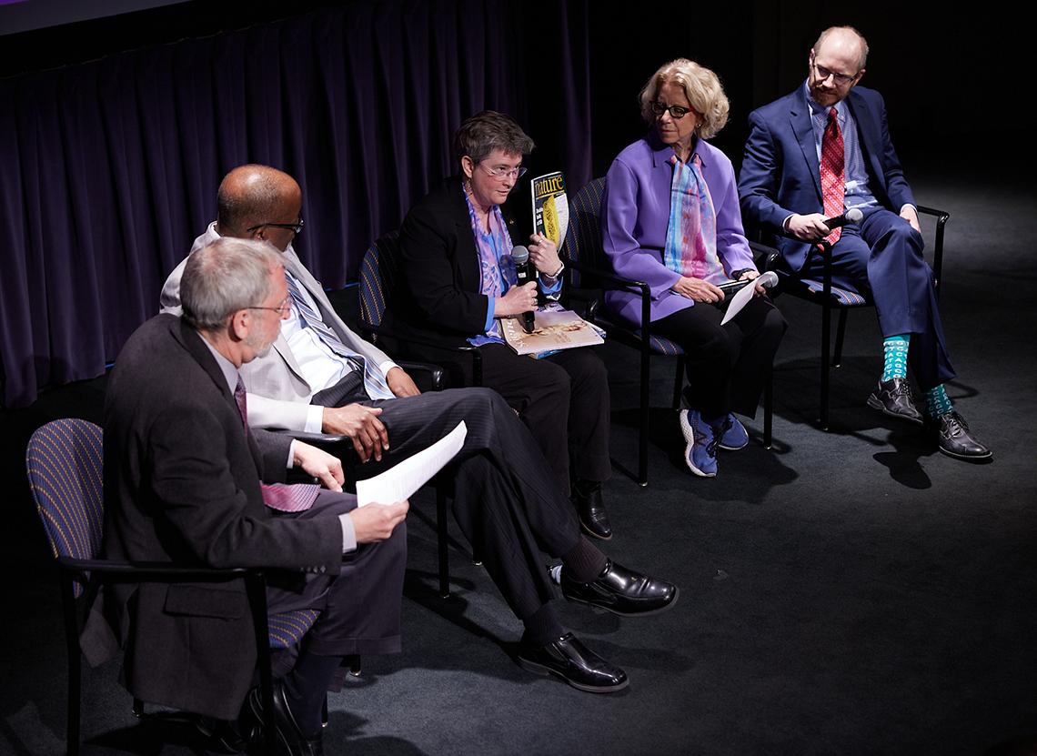 A panel sits on stage, with Dr. Rutter in the middle holding up a copy of Science magazine.