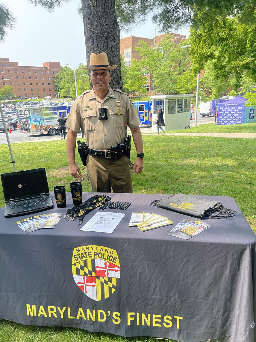 A smiling state trooper stands by info table, with a table covering that reads: Maryland's Finest.