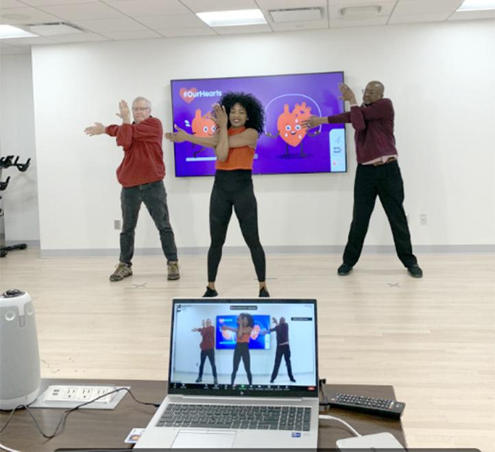 Fitness instructor leads Gibbons and Goff in arm stretches, with laptop on table in front showing virtual recording