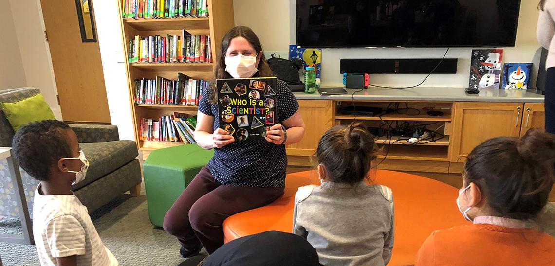 Gehl sits in the Inn lounge, surrounded by books, holding up her book as kids look on.