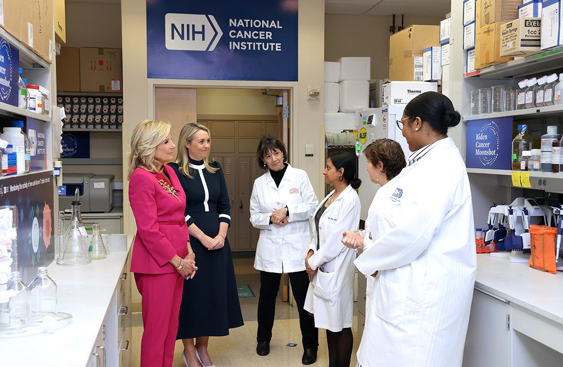 Several women--four in labcoats--chat in a lab environment with NIH NCI logo on wall behind them.