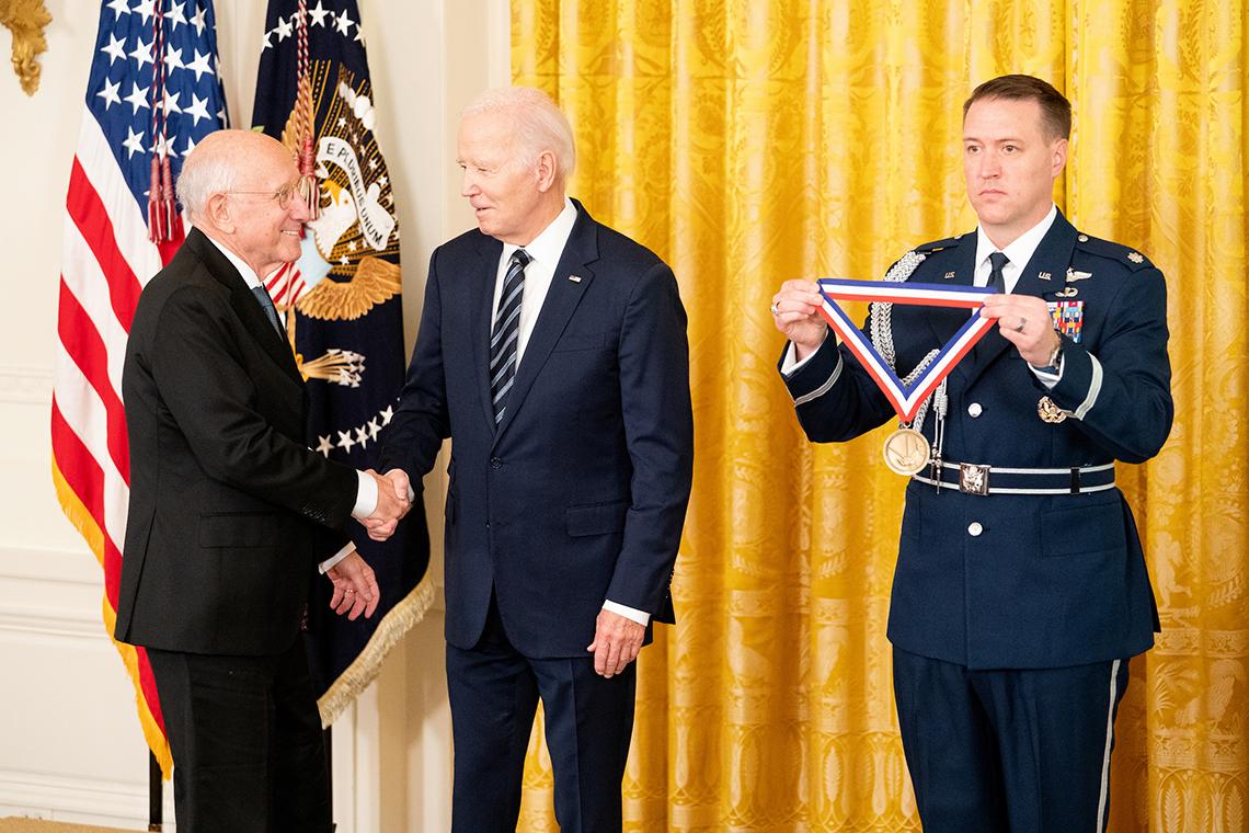 A smiling Rosenberg shakes President Biden's hand as Sargent at Arms holds up the medal, in front of U.S. flag and yellow curtain.