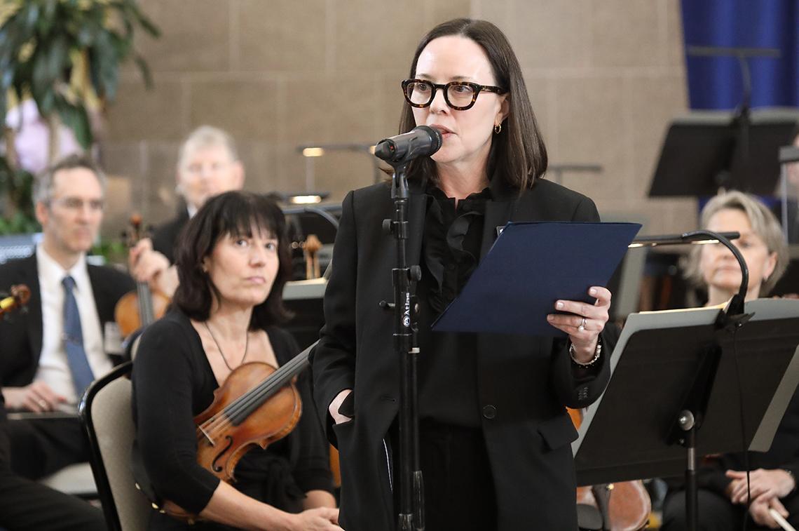Davidson, holding a folder, talks into mic in front of orchestra in the atrium.