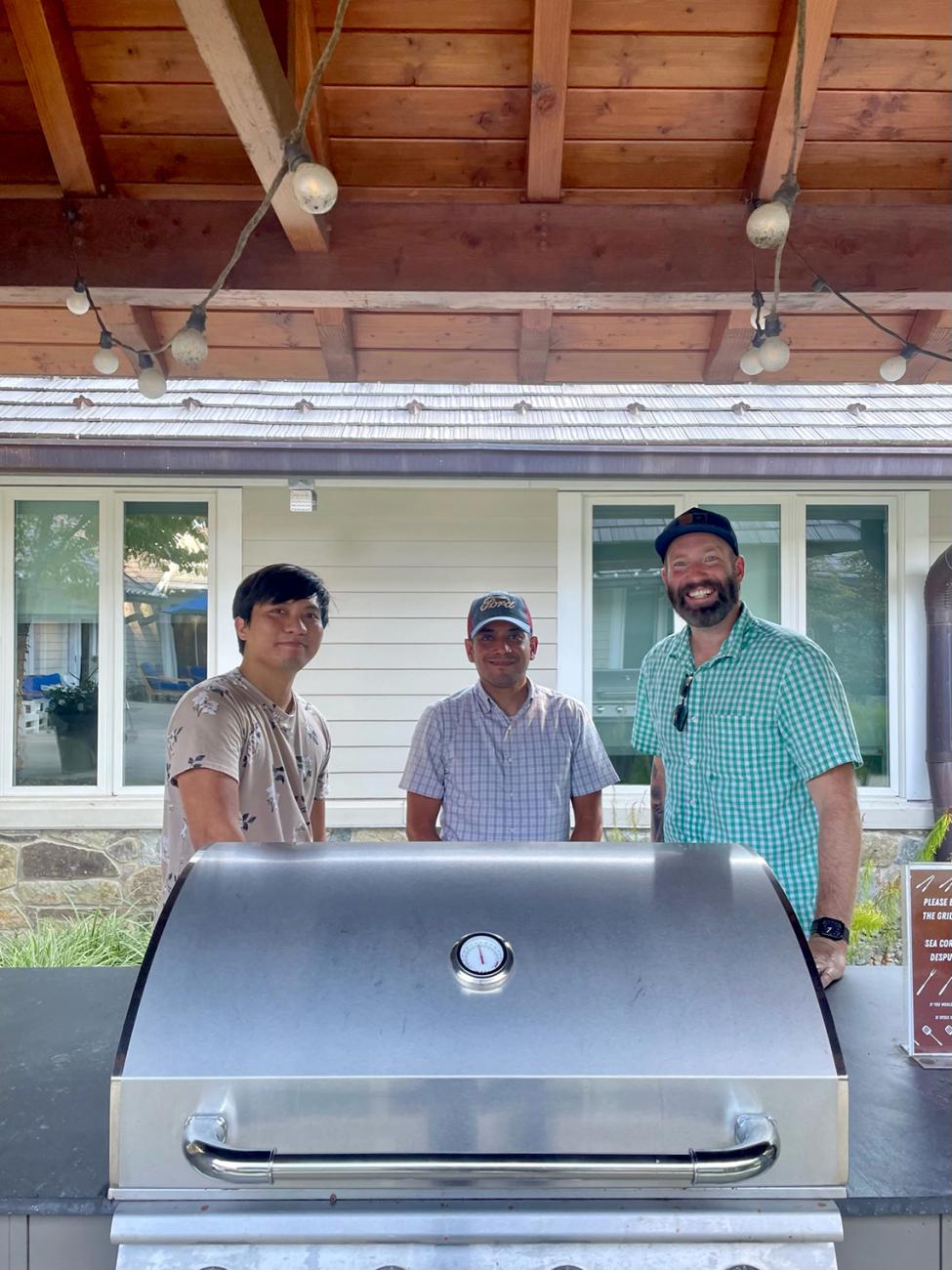 Three men stand behind an outdoor grill.